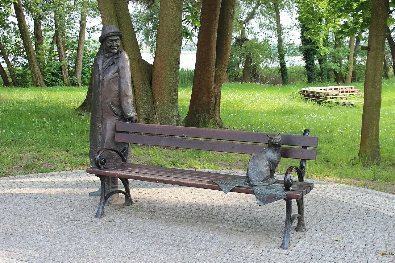 Pomnik mężczyzny w kapeluszu i kota na ławce w parku.