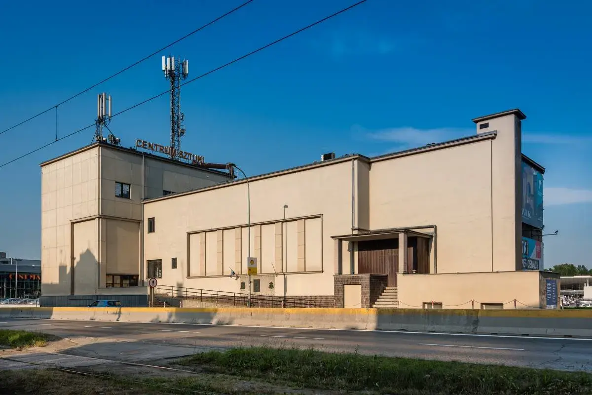 Betonowy budynek z napisem "CENTRUM SZTUKI" na dachu, z wieżami telekomunikacyjnymi. Obok widoczna droga i zieleń.