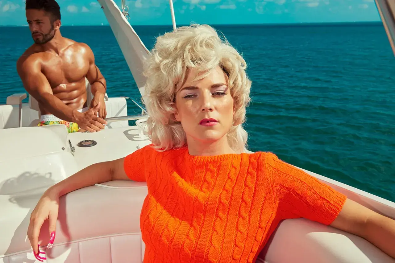 Kourtney Roy in un maglione arancione su una barca, con un uomo muscoloso sullo sfondo, in un'atmosfera estiva.