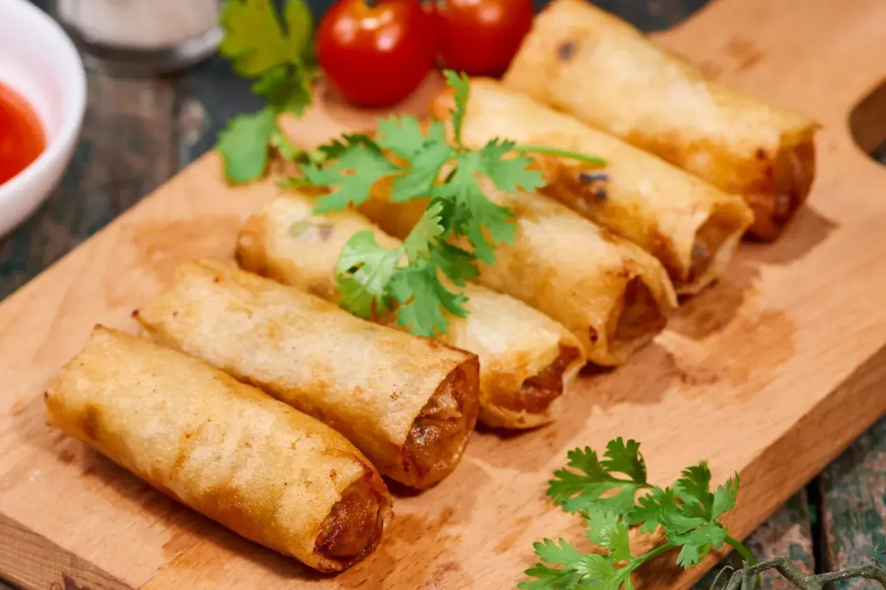 Chrupiące spring rolls na desce, ozdobione kolendrą i pomidorkami. Idealne na przekąskę.