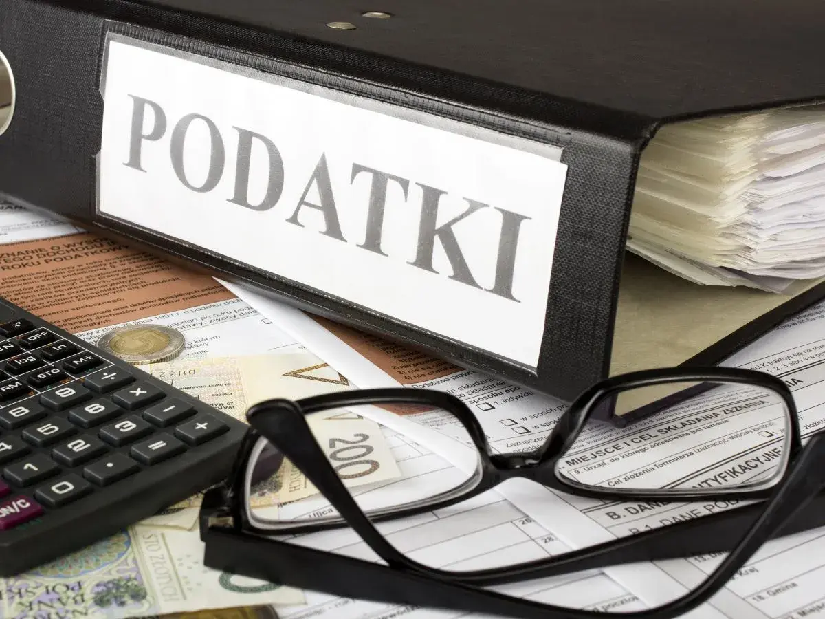 Segregator z napisem "PODATKI", kalkulator, okulary i dokumenty. Obok leży moneta, symbolizująca potencjalną kwotę zmniejszającą podatek.