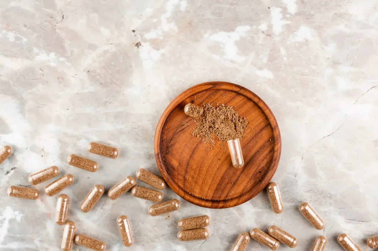 Cápsulas de maca esparcidas sobre un fondo de mármol, con un cuenco de madera que contiene polvo de maca y una cápsula abierta.