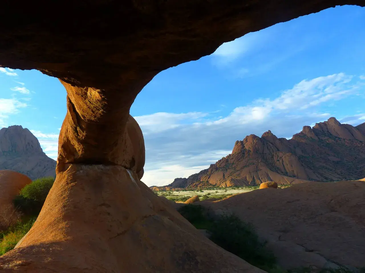 Felsformationen der Spitzkoppe in Namibia, mit einem Felsentor im Vordergrund und zerklüfteten Bergen am Horizont unter blauem Himmel.