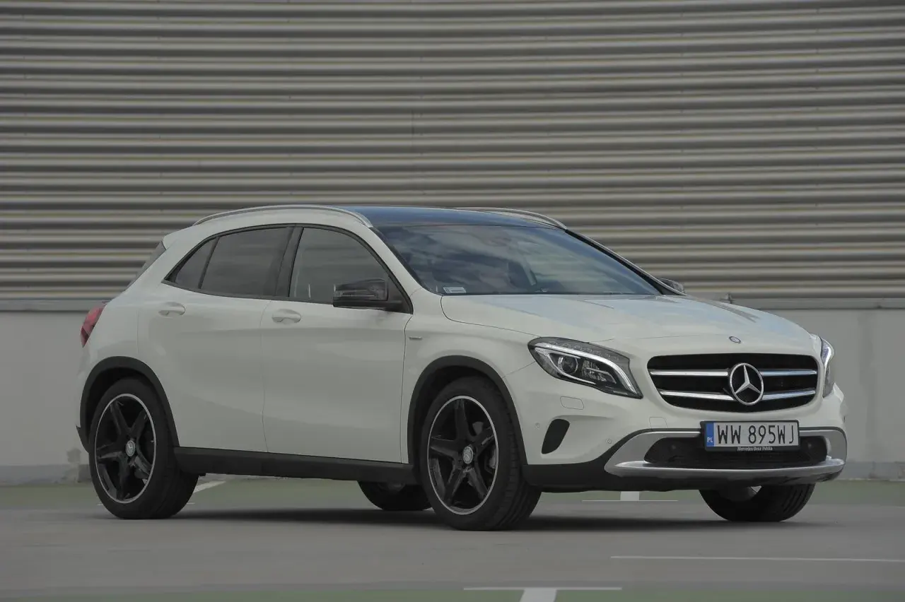 Biały Mercedes GLA na tle szarej, falistej ściany. Zbliżenie na przód auta, widoczne logo i tablica rejestracyjna. Idealny do miasta, poznaj jego mercedes gla wymiary.