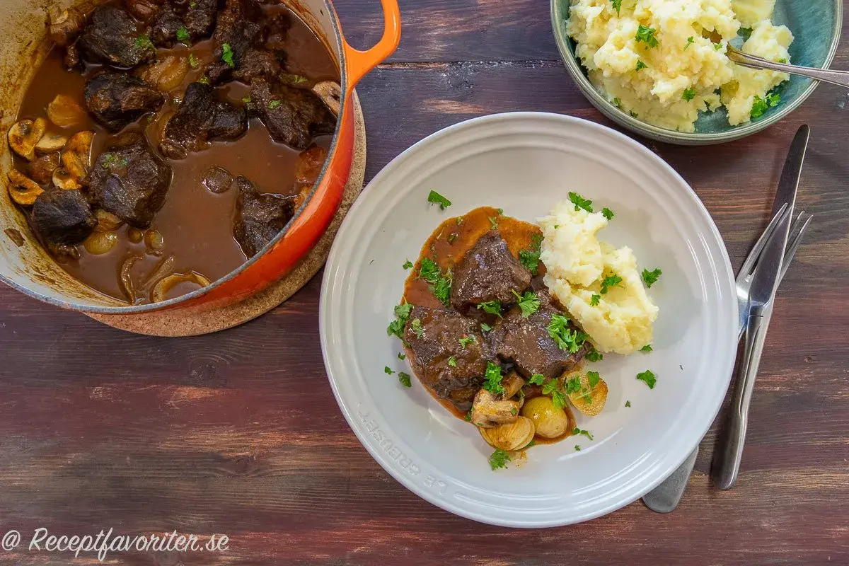 A Dutch oven filled with braised beef and mushrooms, next to a plate of beef with mashed potatoes and a bowl of mashed potatoes.