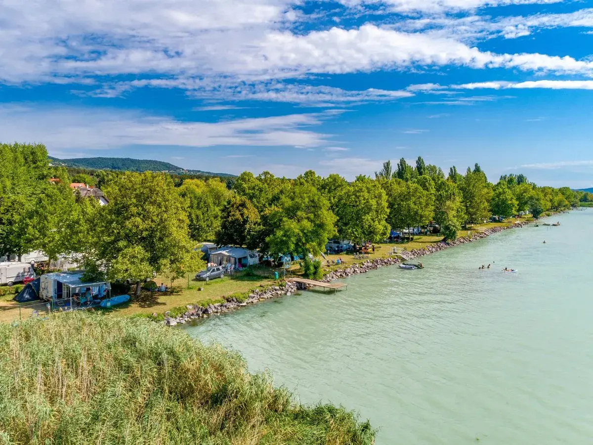 Słoneczny dzień na kempingu Balaton. Przyczepy kempingowe i namioty nad turkusową wodą, ludzie pływają i relaksują się.