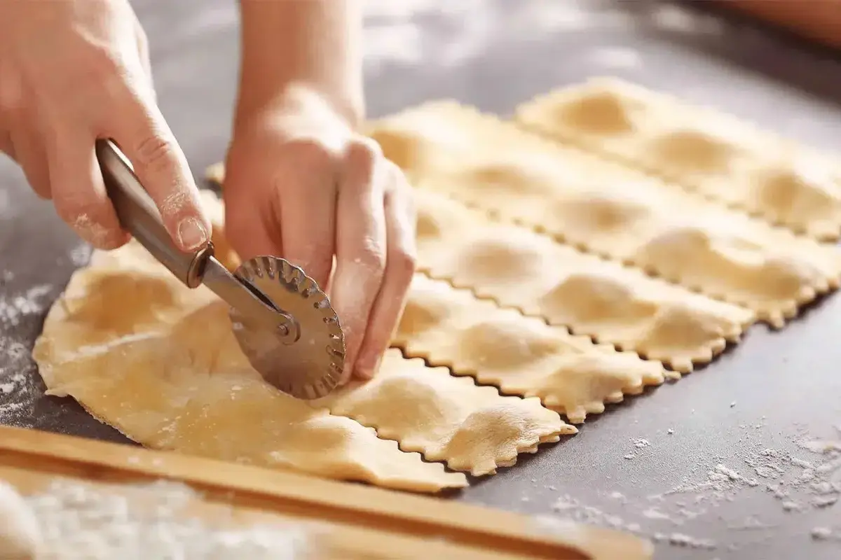 Préparation d'une recette de ravioli maison. Des mains découpent la pâte avec une roulette dentelée, formant des carrés prêts à être farcis.