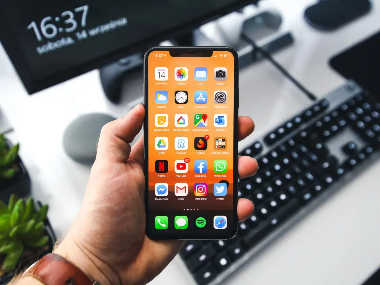 Dłoń trzyma iPhone'a z widocznymi ikonami aplikacji. Na ekranie komputera w tle widnieje godzina 16:37. Zastanawiasz się, jak wyłączyć iPhone'a?