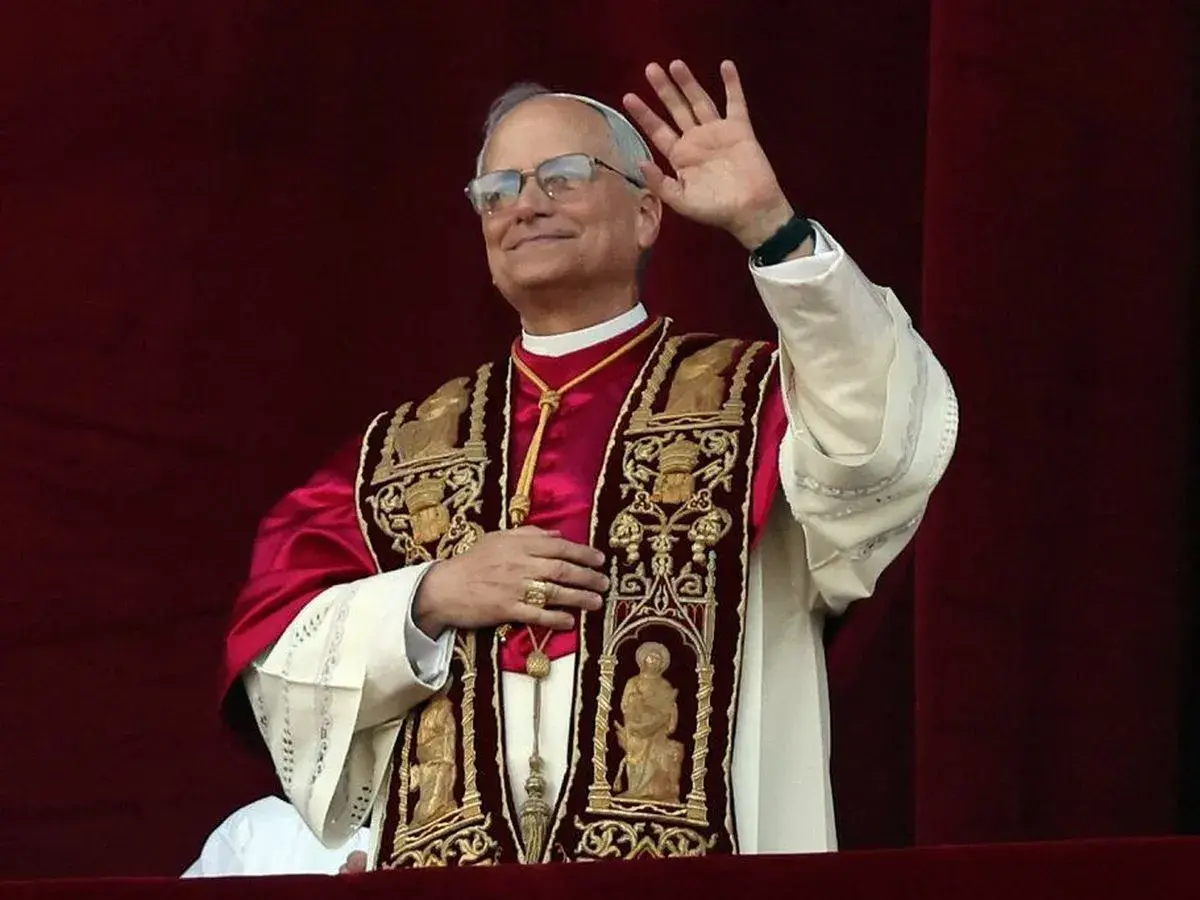 Papież w bogato zdobionej szacie pozdrawia wiernych. Jego uśmiech i gest ręki budzą nadzieję, a jego strój przypomina o tym, skąd pochodzi papież i jego dziedzictwo.