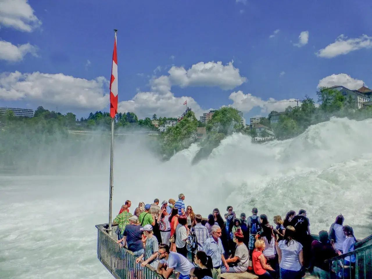 Menschen bestaunen den tosenden Rheinfall. Eine Schweizer Fahne weht im Wind. Der **Rheinfall Eintritt** ist ein beeindruckendes Naturschauspiel.