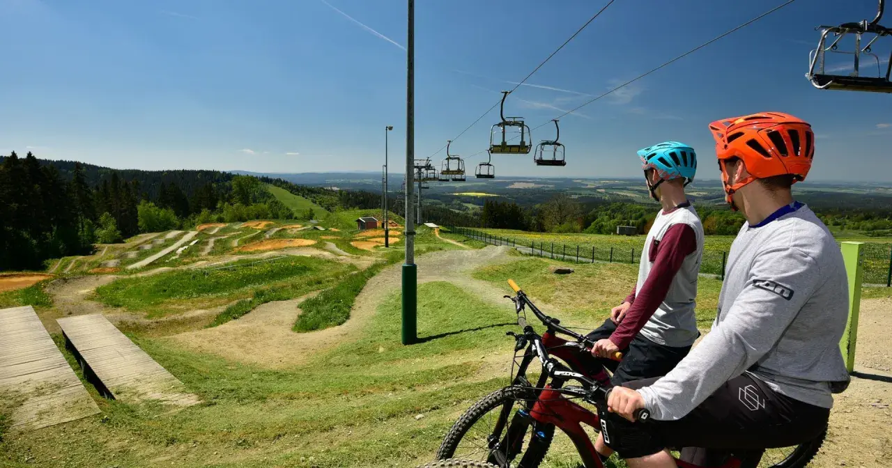 Zwei Mountainbiker mit Helmen blicken auf einen aufregenden mountainbike trail mit Sprüngen und Wellen. Eine Seilbahn schwebt darüber.