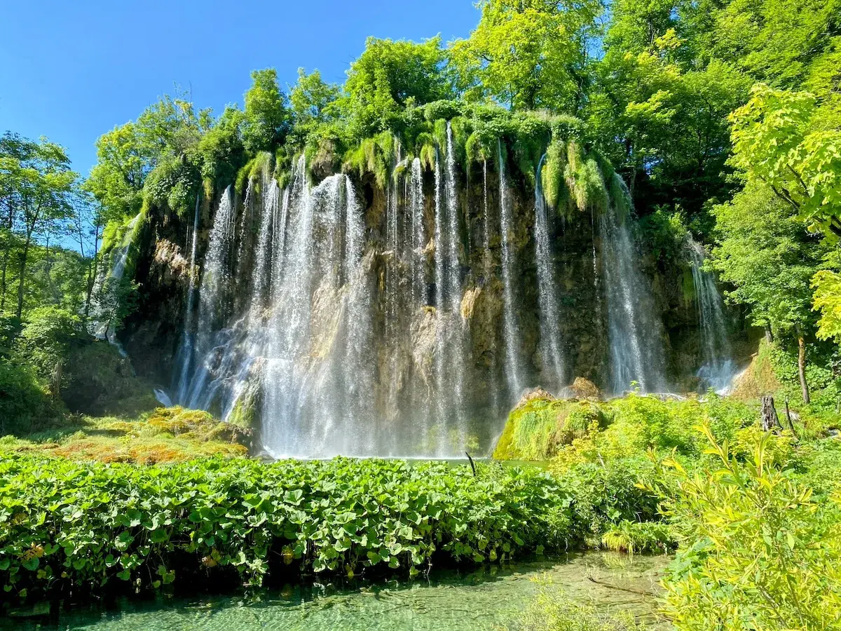 A stunning waterfall cascades down a rocky cliff, surrounded by lush green foliage. Perfect for Croatia hiking trips, this scene captures the natural beauty of Plitvice Lakes National Park.