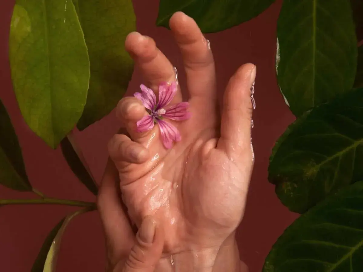 Main délicate tenant une fleur rose, entourée de feuilles vertes. Une image évoquant la sérénité et les bienfaits du Jin Shin Jyutsu.