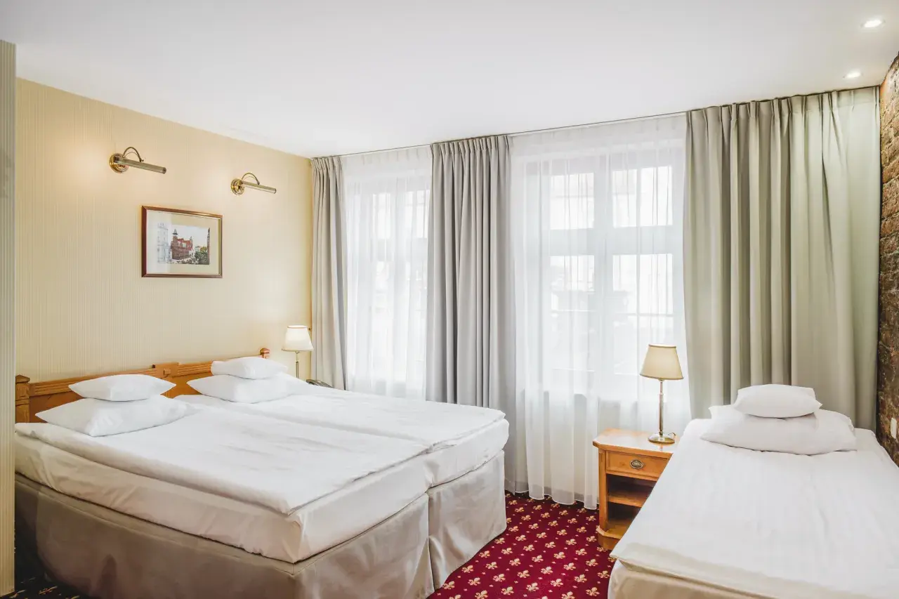 Pokój hotelowy z trzema łóżkami, idealny na wieczór panieński. Jasne ściany, zasłony i czerwony dywan tworzą przytulną atmosferę.