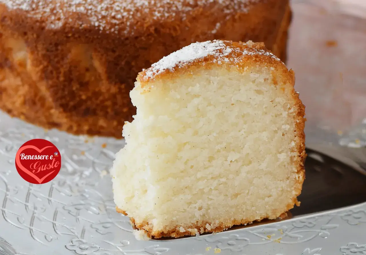 Una fetta di torta senza glutine, soffice e dorata, cosparsa di zucchero a velo, su un piatto trasparente.