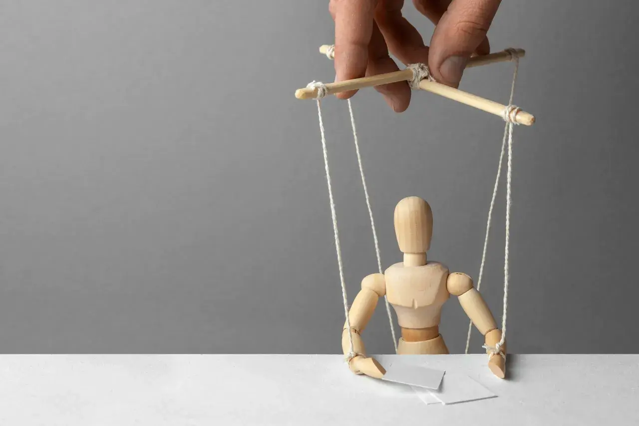 Drewniany ludzik-marionetka, sterowany przez ludzką dłoń, symbolizuje podżeganie i manipulację.