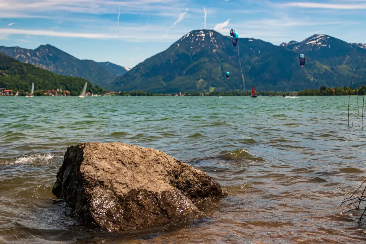 Kitesurfer tanzen über das Wasser am Bad Wiessee, während Segelboote und Berge im Hintergrund ruhen.