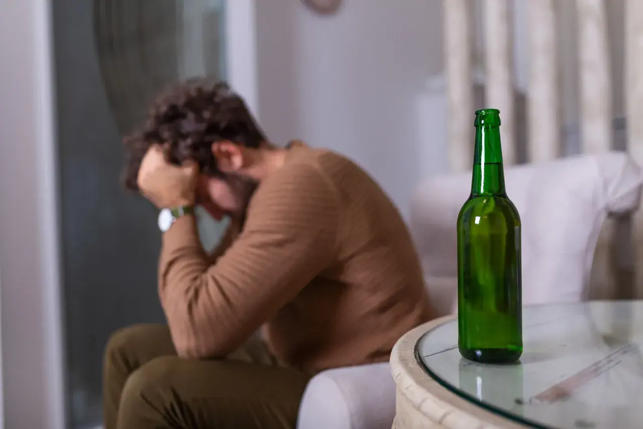 Mężczyzna z głową w dłoniach siedzi obok pustej butelki piwa. Może to symbolizować uczulenie na alkohol lub jego nadużywanie.