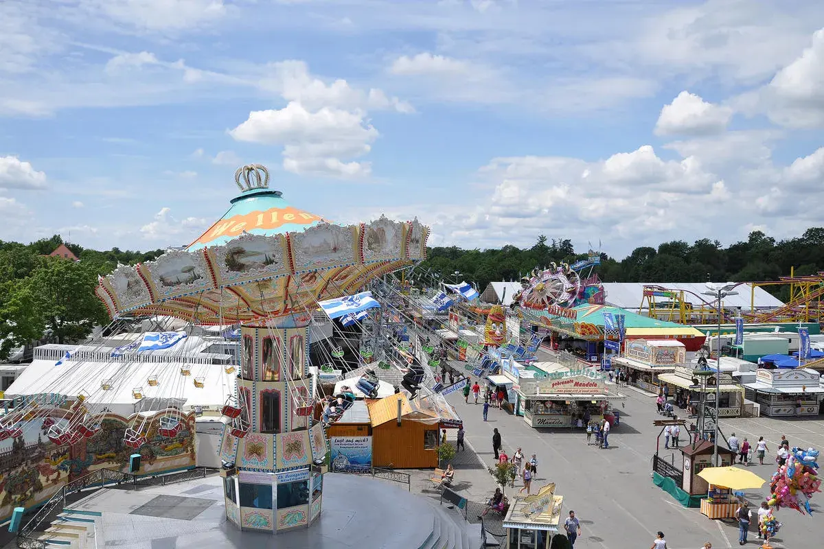 Buntes Treiben auf dem Volksfest Ingolstadt mit Kettenkarussell, Fahrgeschäften und Buden unter blauem Himmel mit Wolken.