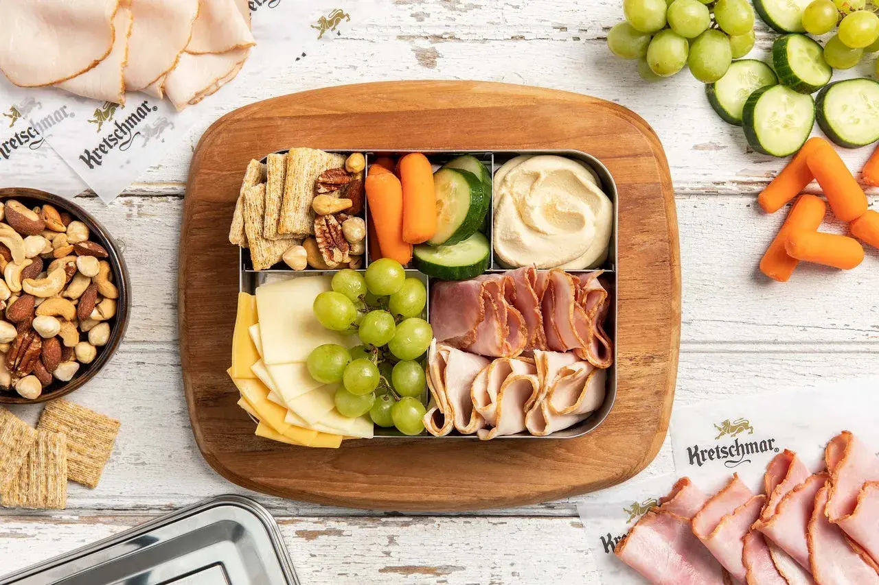 A bento box with cheese, grapes, crackers, nuts, hummus, carrots, and cucumber. Perfect for when are bento boxes eaten cold.
