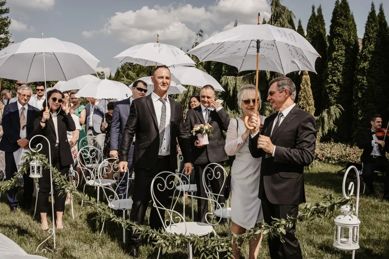 Wesele w kwietniu pod białymi parasolami. Goście w eleganckich strojach czekają na ceremonię na trawie, wśród zieleni i ozdobnych krzeseł.