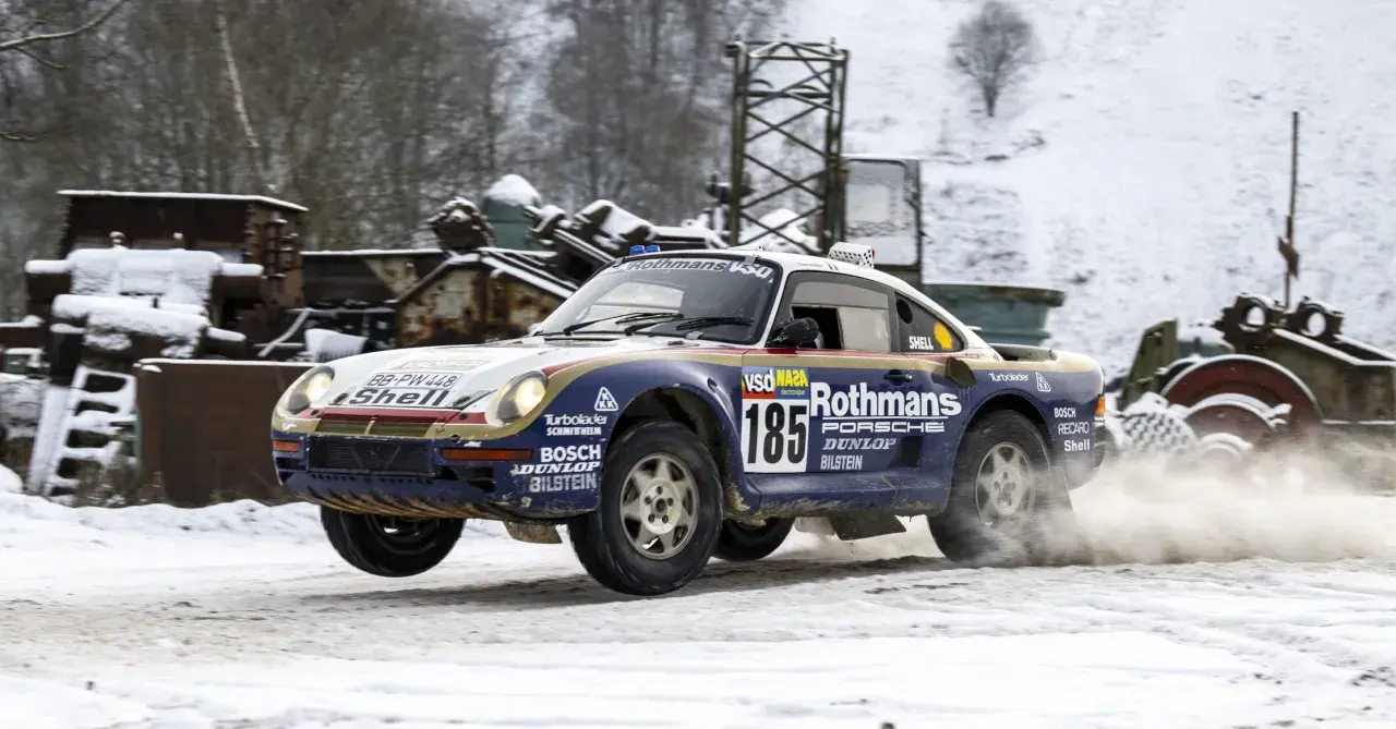 Porsche 959 w barwach Rothmans, z numerem 185, pokonuje śnieżny odcinek rajdu, wzbijając tumany śniegu.