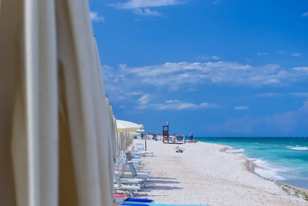 File di ombrelloni bianchi aprono la vista sulla spiaggia di Marotta, con lettini pronti e il mare turchese sotto un cielo azzurro.