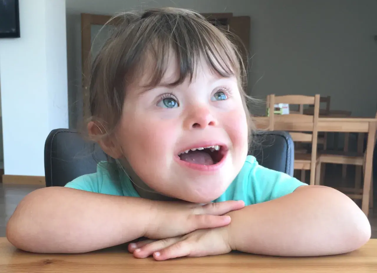 Una bambina down con occhi azzurri e un sorriso radioso, appoggiata al tavolo con le braccia incrociate.