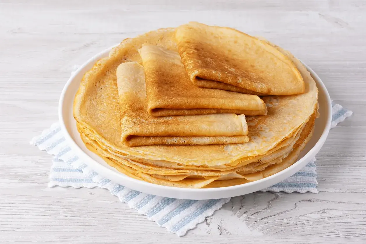 Pile de crêpes dorées, certaines pliées, sur une assiette blanche. Un délice sans oeuf pour un petit-déjeuner léger.