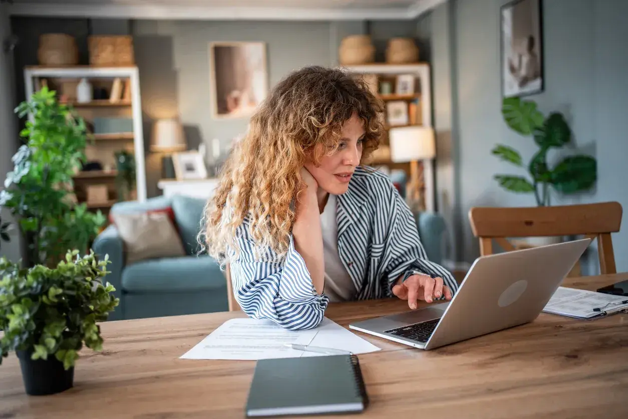 Donna con capelli ricci lavora al computer, documenti sul tavolo. Forse sta cercando informazioni su un mutuo con garante genitore per realizzare un sogno.