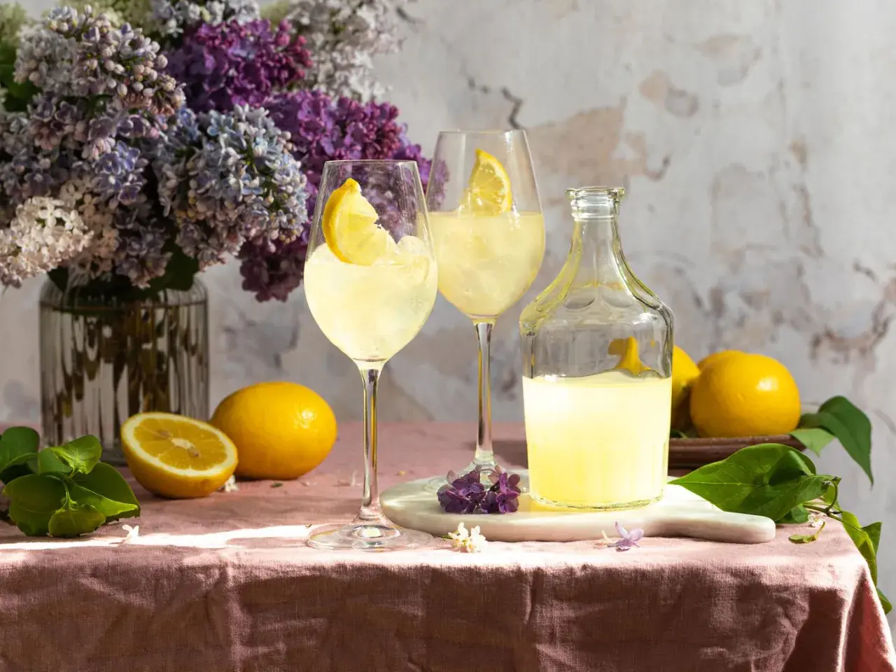 Zwei Gläser Limoncello Spritz mit Zitrone, eine Flasche Limoncello, Zitronen und Flieder auf rosafarbenem Tischtuch.