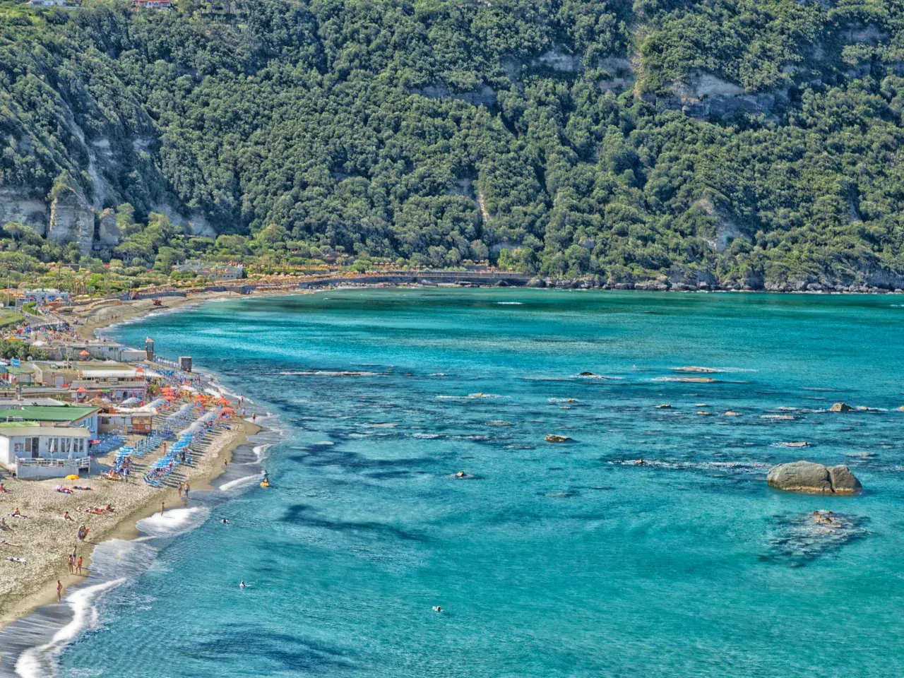 La spiaggia di Citara, con le sue acque cristalline e la rigogliosa vegetazione alle spalle, accoglie bagnanti e ombrelloni colorati.