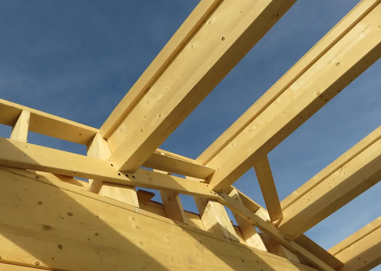 Structure en bois d'une charpente fermette sous un ciel bleu. Les poutres de bois clair forment un motif géométrique complexe.