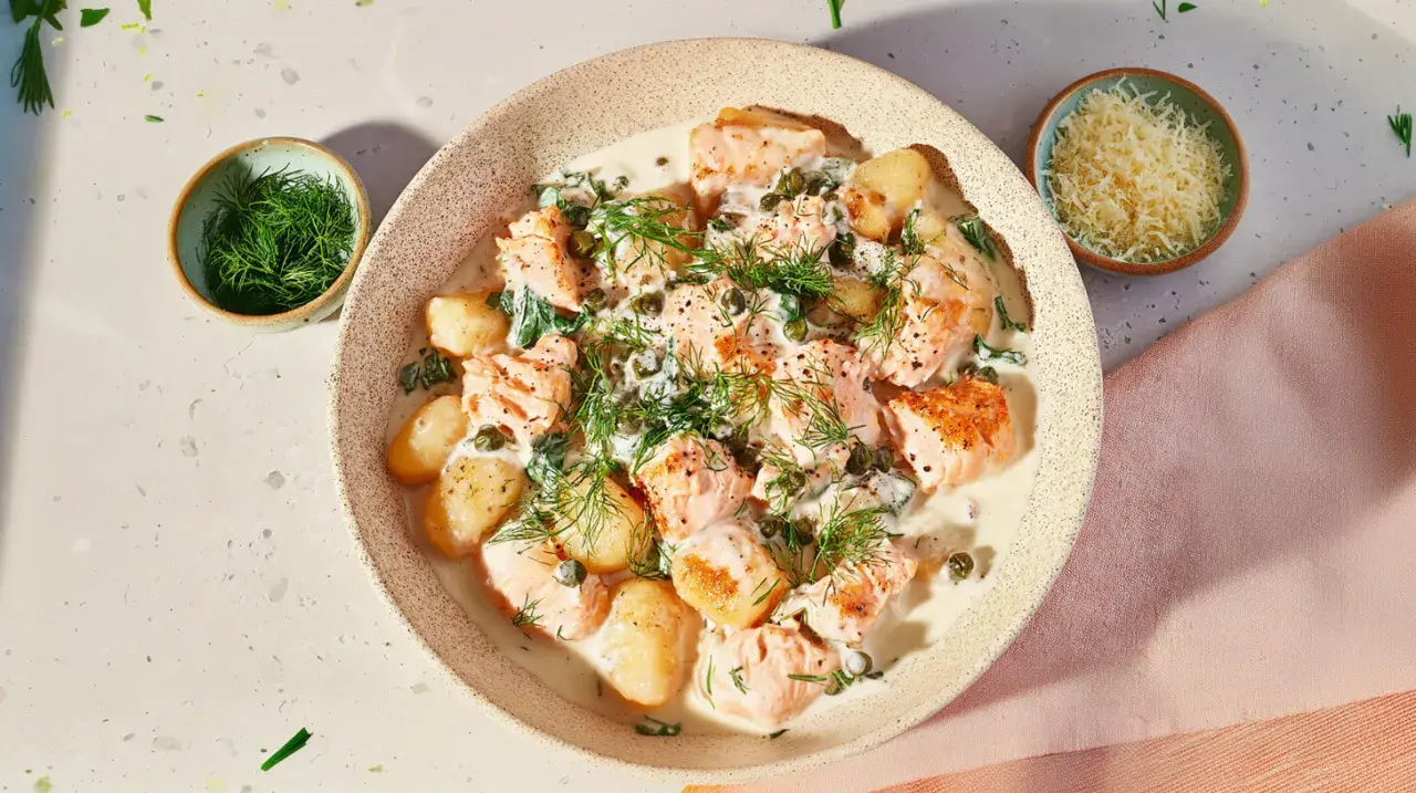 Pyszne gnocchi z łososiem w kremowym sosie, udekorowane koperkiem i kaparami. Obok miseczka z tartym parmezanem.