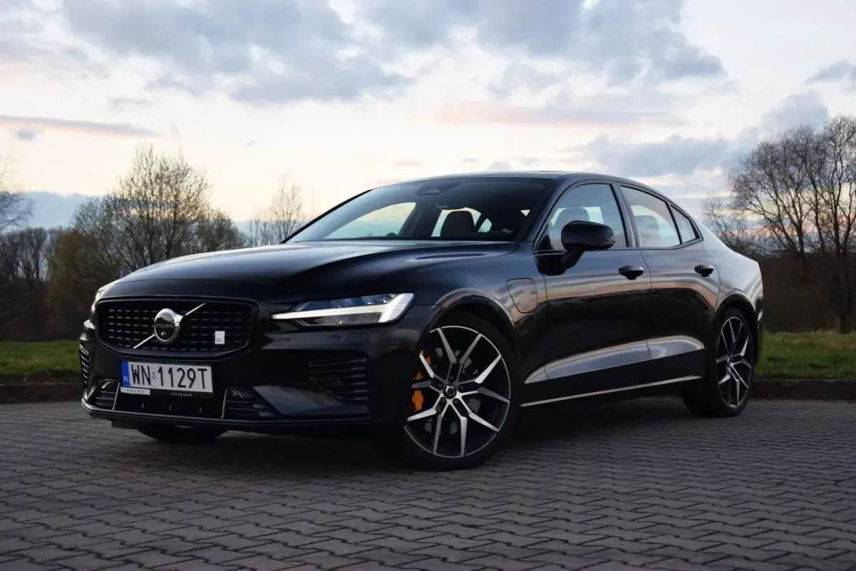 Czarny Volvo S60 T8 na tle zachodzącego słońca i drzew. Elegancki sedan z charakterystycznym grillem i felgami.