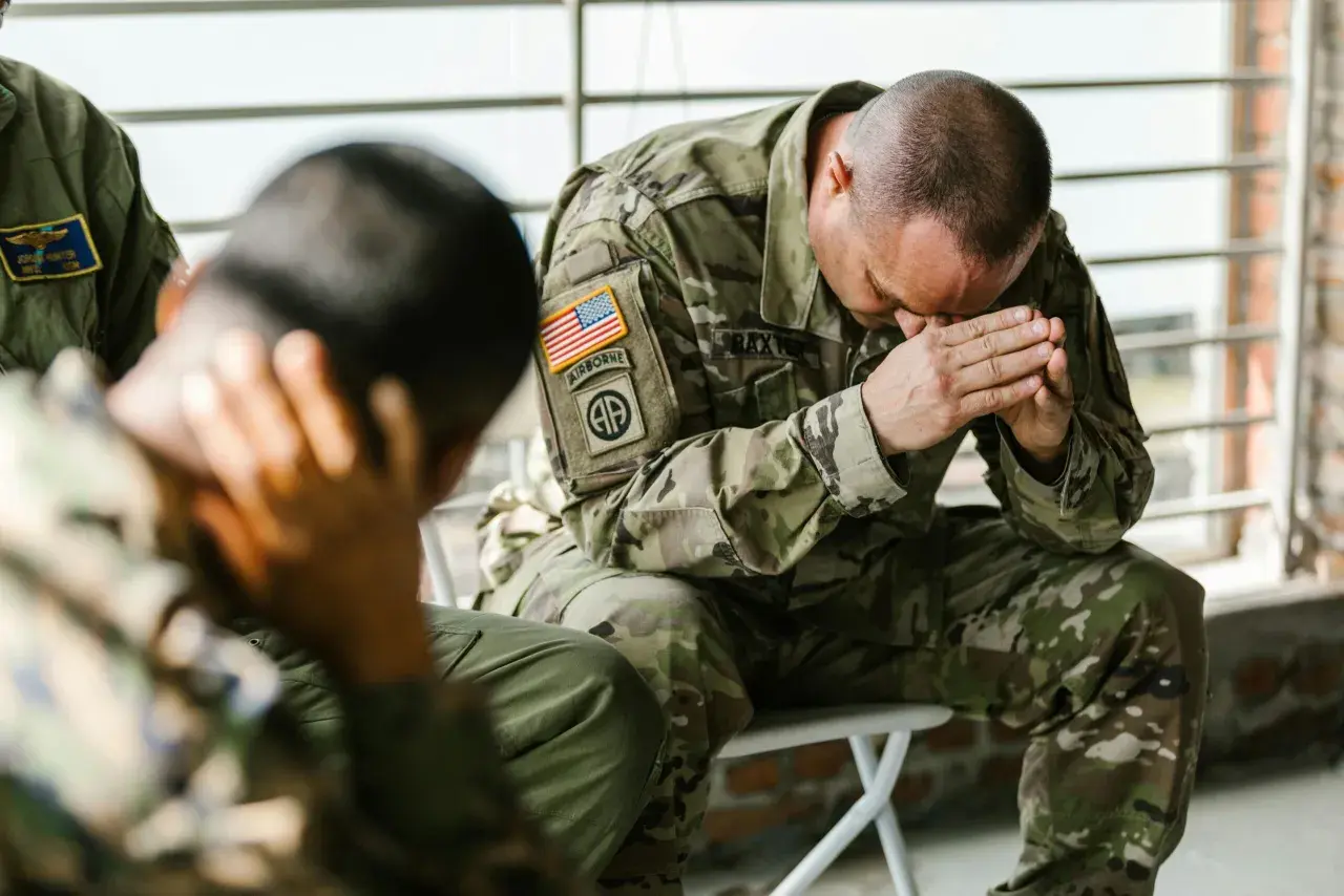 Żołnierze w mundurach siedzą, jeden z nich z rękami na twarzy, co może symbolizować walkę z PTSD.