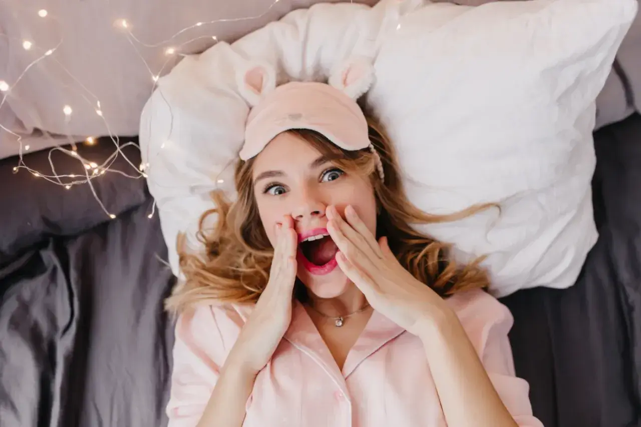 En tjej med sovmask och rosa pyjamas ligger i sängen, redo för att sova med sminket kvar. Hon ser överraskad ut.