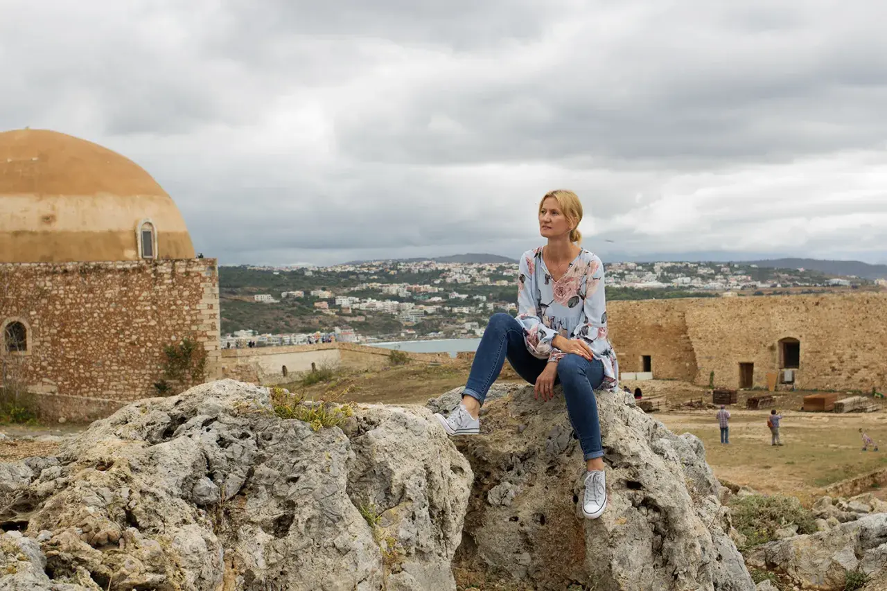 Kobieta siedzi na skale, podziwiając widok miasta. Kreta pogoda październik, chmury na niebie.