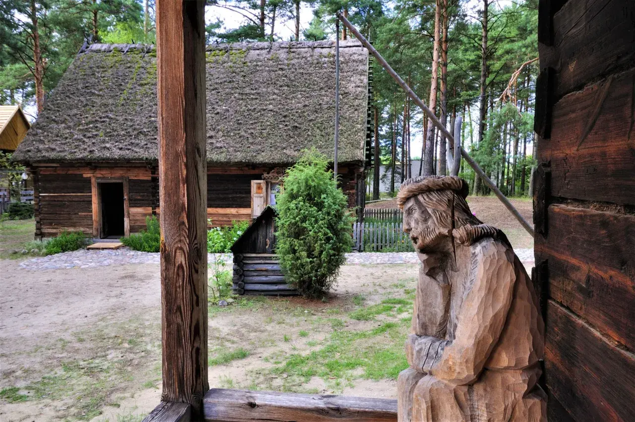 Drewniana rzeźba postaci, tradycyjna chata z mchem na dachu i zielone drzewa tworzą klimatyczne **kadzidło atrakcje** w skansenie.