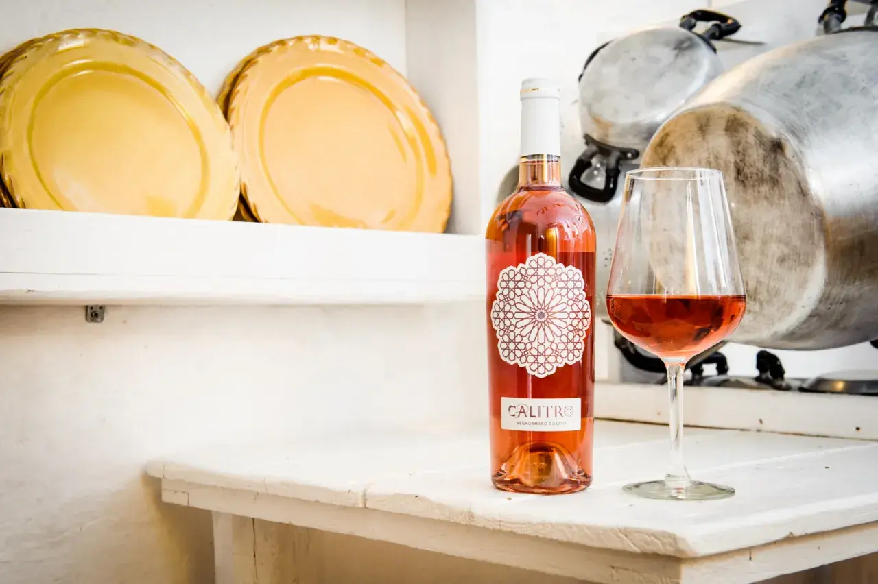 Un bicchiere e una bottiglia di negroamaro rosato Calitro su un tavolo bianco, con piatti gialli e pentole sullo sfondo.