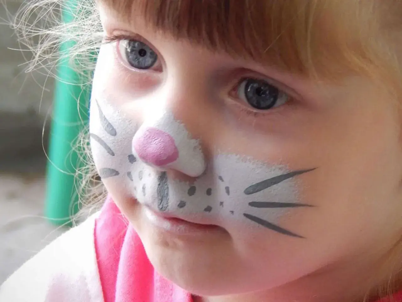 Ein kleines Mädchen mit blauem Auge und Kinderschminken als Tier. Die Nase ist rosa, die Wangen mit Punkten und Schnurrhaaren bemalt.