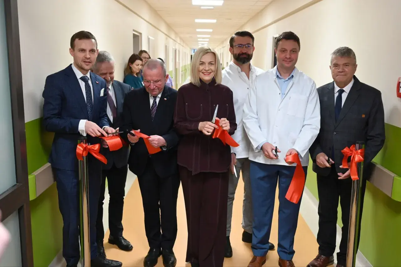 Grupa osób w garniturach i kitlach przecina czerwone wstęgi w szpitalnym korytarzu.