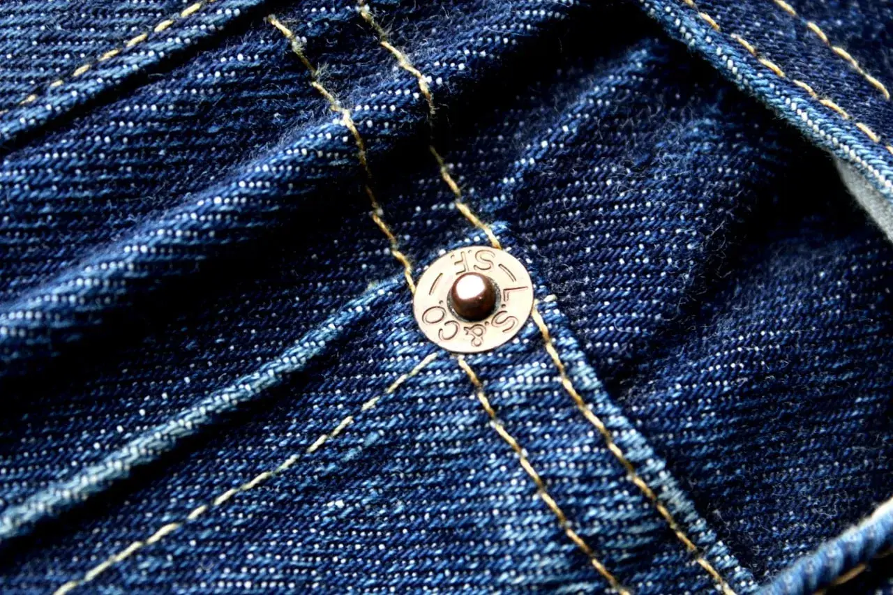 Close-up of dark blue denim with yellow stitching. A copper rivet, marked "S.F. & CO. L.S.", is visible, a testament to the durability of these jeans.