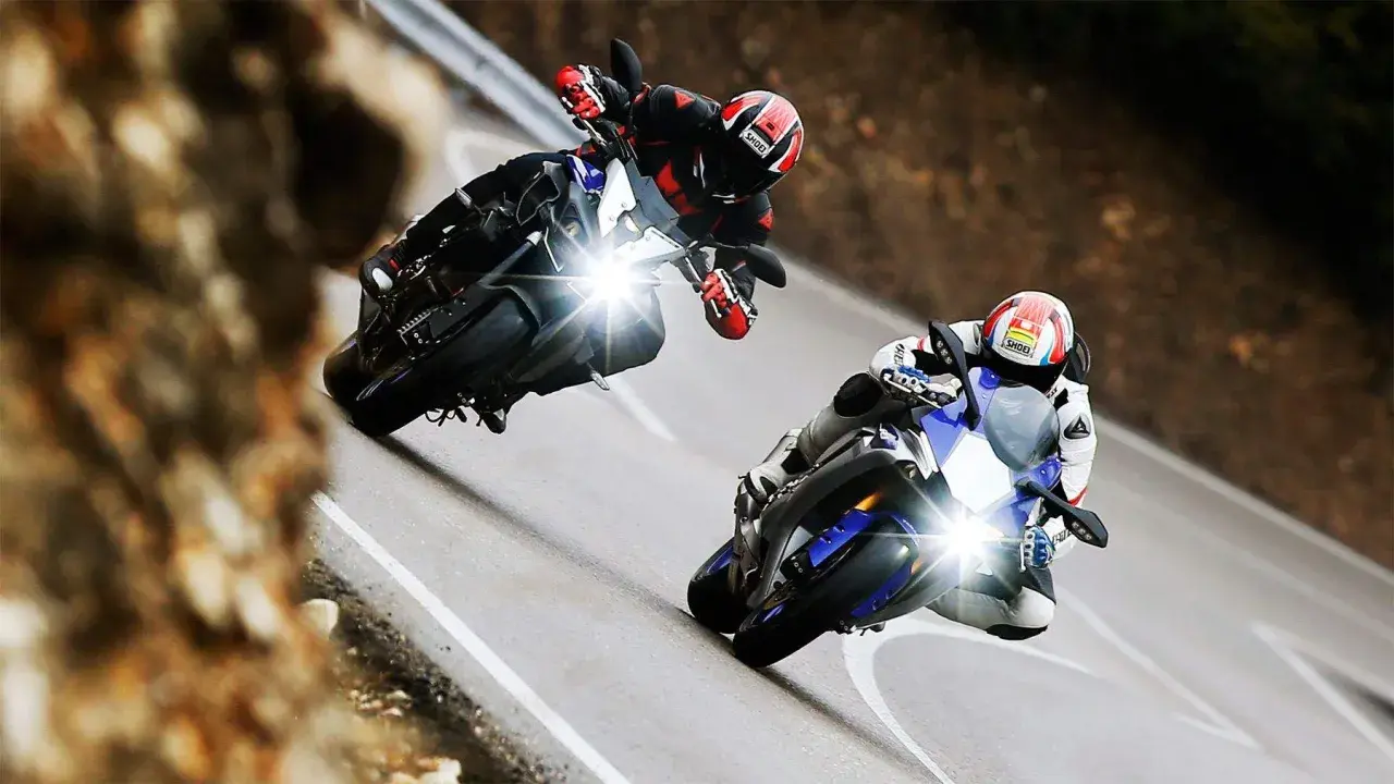 Dos pilotos compiten en una carretera sinuosa, uno en una Yamaha mas rápida, inclinándose en una curva cerrada.