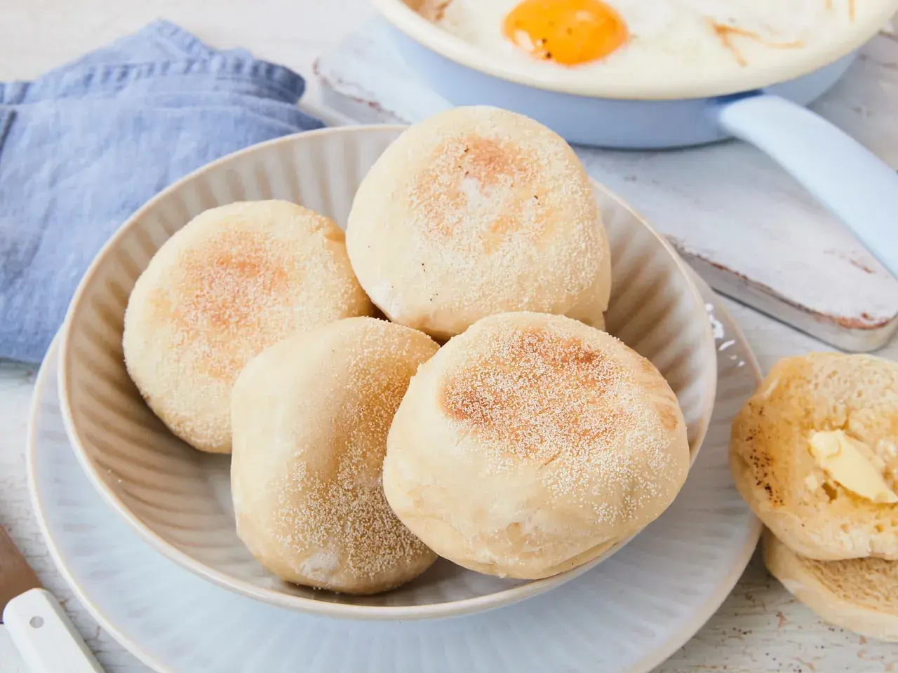 Ein Teller mit vier goldbraunen englischen Muffins, daneben ein halber, mit Butter bestrichener Muffin. Ein Spiegelei ist im Hintergrund zu sehen.