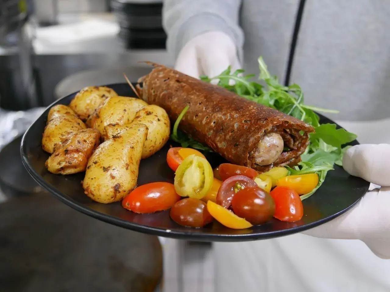 Une galette saucisse roulée, servie avec des pommes de terre rôties et des tomates cerises colorées, sur une assiette noire.