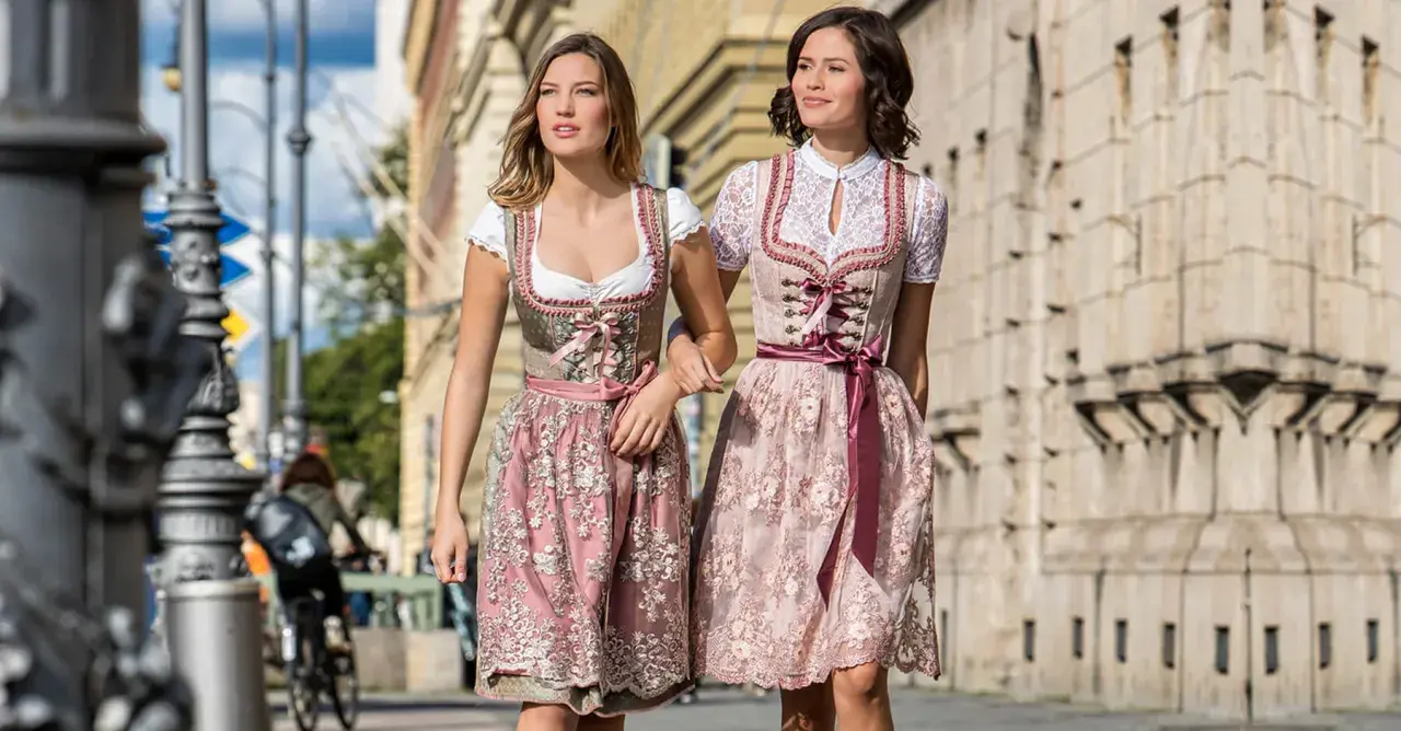 Zwei Frauen in Dirndln ohne Schnürung spazieren. Die Kleider sind in Rosa- und Beigetönen gehalten und mit zarten Stickereien verziert.