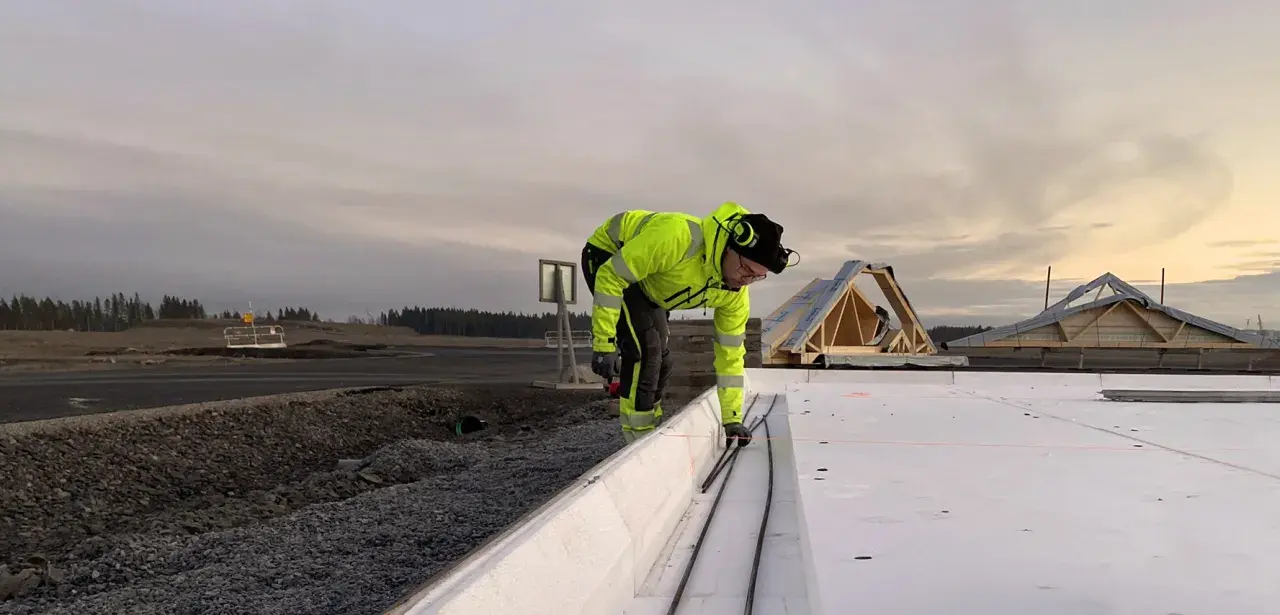 Arbetare i varselkläder förbereder armering för betongsten grund. Takstolar syns i bakgrunden.