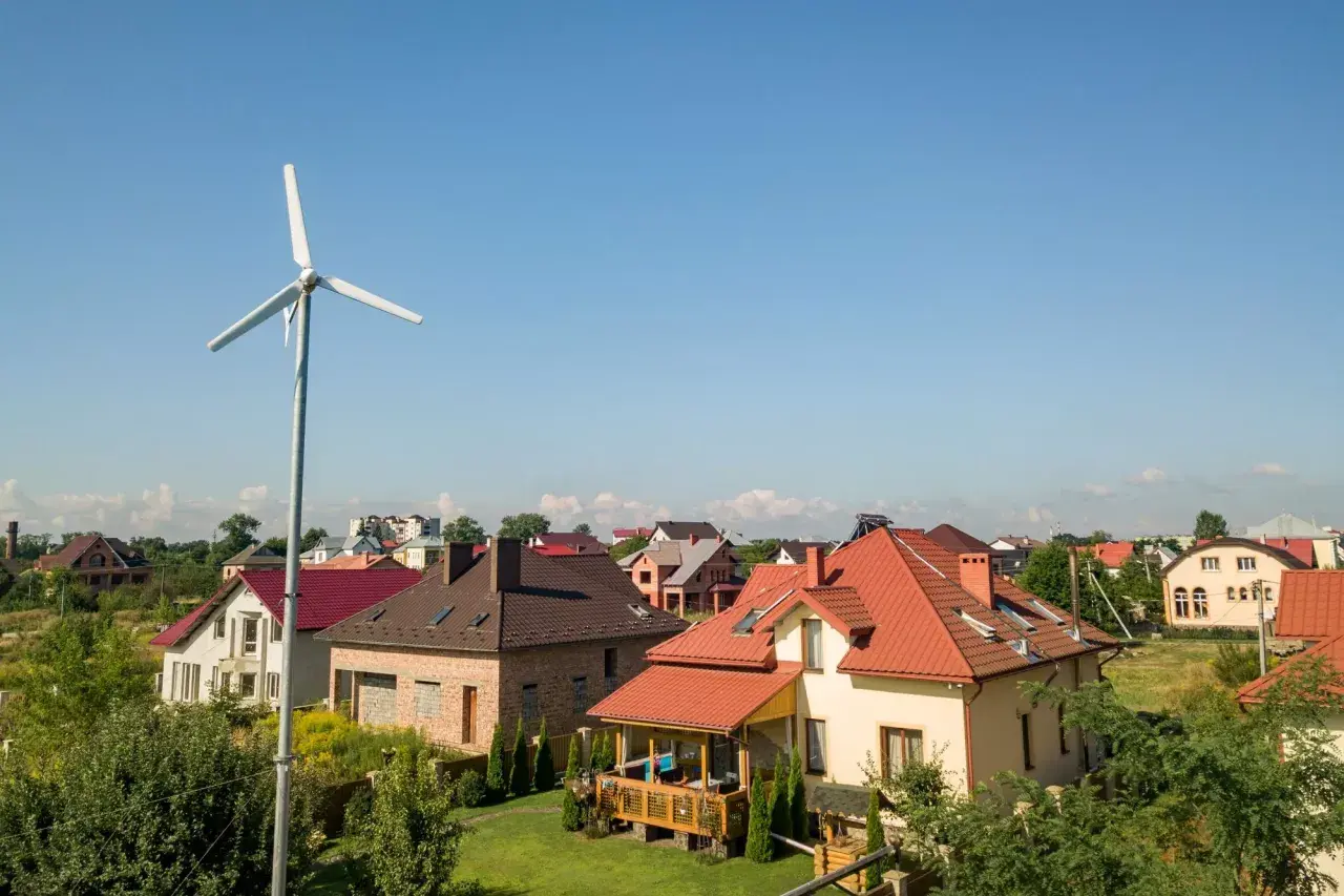 Biała turbina wiatrowa na tle nieba i domów jednorodzinnych.