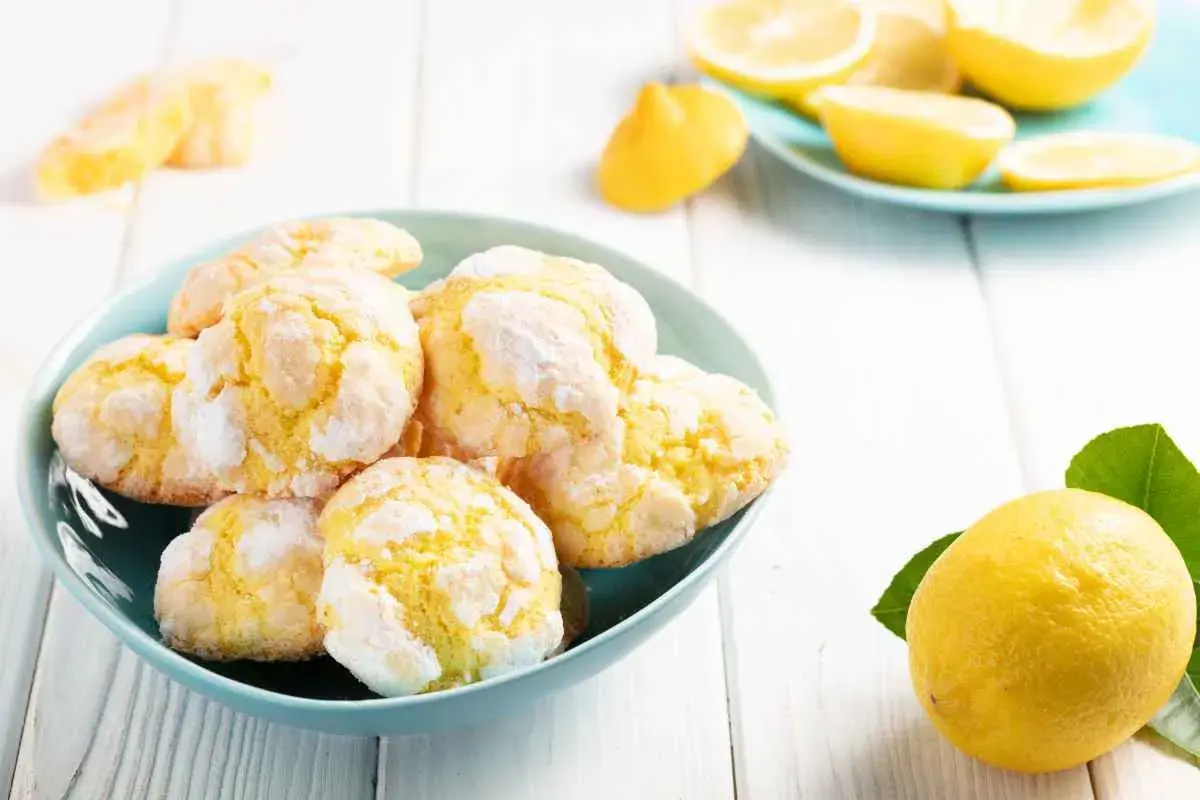 Biscotti al limone, morbidi e profumati, in una ciotola azzurra, con limoni freschi e spicchi su un piattino sullo sfondo.